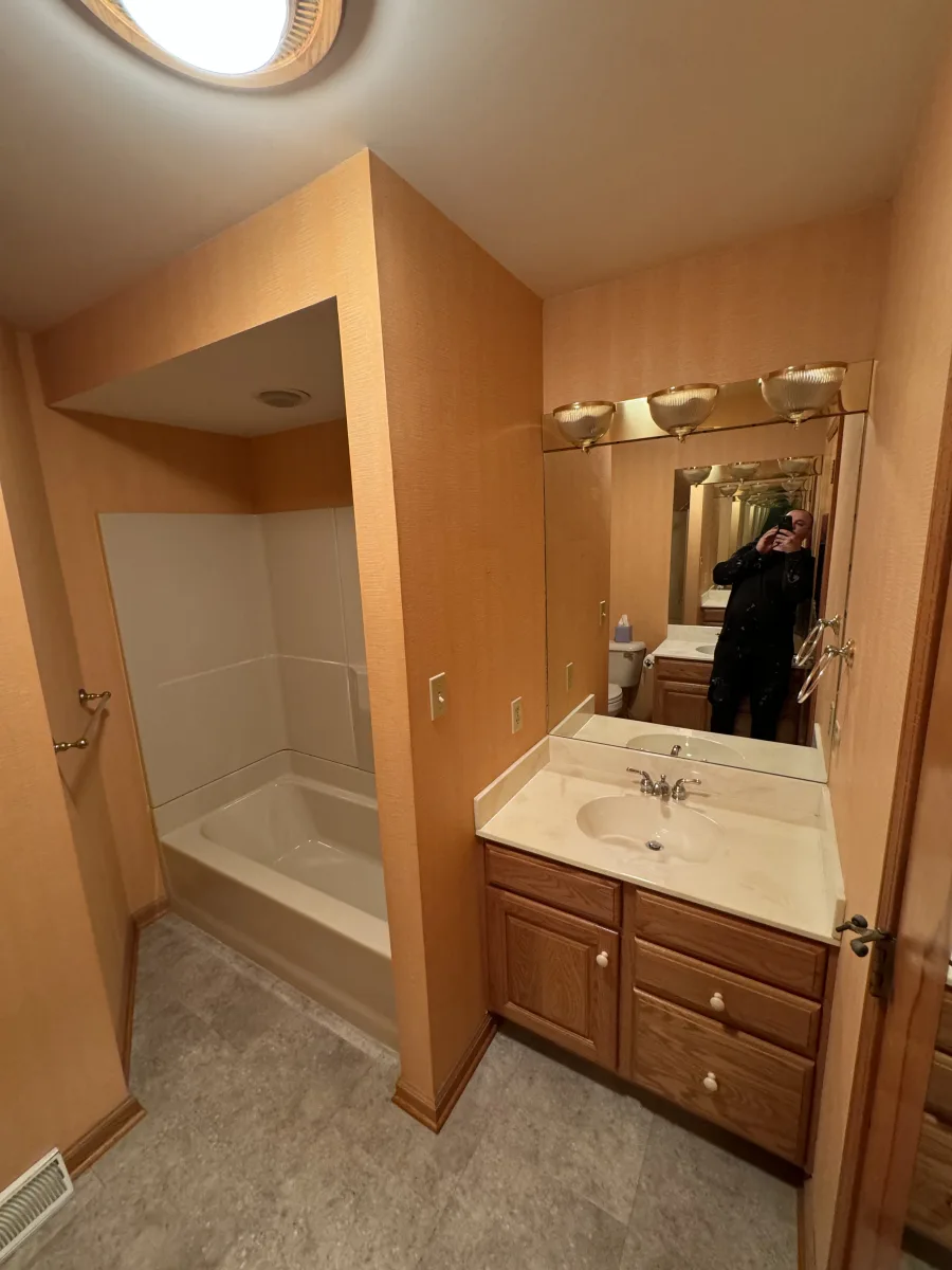 Columbus guest bathroom before renovation with orange wallpaper, wood vanity, white tub, gold lights, and tile.